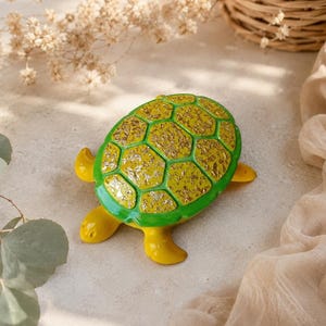 Op de afbeelding: Een decoratief schildpadbeeldje met een felgele kop en ledematen, en een schild met een levendige groene rand en een centrum gevuld met gestructureerde bladgoudvlokken. De schildpad is afgebeeld in een bovenaanzicht dat het schild toont.