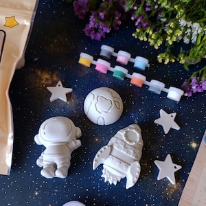 May include: A space-themed craft kit with plaster figures of an astronaut, rocket, planet, and stars. The kit includes small paint pots in various colours and a paintbrush. The items are arranged on a dark blue surface with a starry pattern.