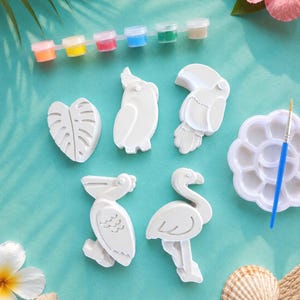 Può includere: Un kit artigianale con forme di uccelli e foglie in gesso bianco, vasetti di vernice, un pennello e una tavolozza. Il kit include un tucano, un pappagallo, un pellicano e un fenicottero. Lo sfondo è azzurro con fiori tropicali e conchiglie.
