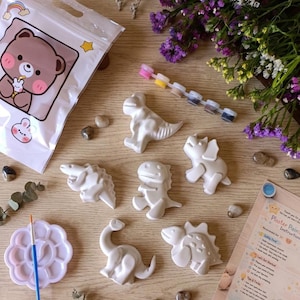 Könnte beinhalten: Ein Bastelset mit sechs weißen Dinosaurierfiguren aus Gips, einer Farbpalette, Farbtuben und einem Pinsel. Ein Beutel mit einer Bärenillustration und Anweisungen sind ebenfalls enthalten. Das Set ist auf einer Holzoberfläche angeordnet.