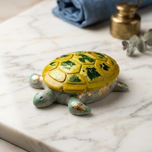 May include: A ceramic turtle ornament with a yellow and green shell, green head and limbs, and gold detailing. The turtle is on a marble surface. Decorative item.