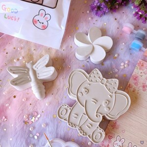 Puede incluir: Colección de figuras de cerámica sin pintar, incluyendo una libélula, una flor y un elefante con un lazo. Las figuras están dispuestas sobre una superficie rosa y morada con pintura y un pincel. También se ve una bolsa con el texto "Good Luck!".