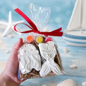 Könnte beinhalten: Ein klarer Zellophanbeutel, gebunden mit einem roten Band, enthält zwei weiße Kekse mit Meeresmotiven und bunte Bonbons. Einer ist eine Qualle, der andere eine Meerjungfrauenflosse. Im Hintergrund sind ein Spielzeugsegelschiff und Muscheln.