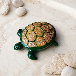 Könnte beinhalten: Dekorative Schildkrötenfigur mit grünem Körper und einem Panzer mit goldgelbem Mosaikmuster. Die Figur liegt auf einer hellen Oberfläche, zusammen mit kleinen, runden Steinen und korallenartigen Objekten.