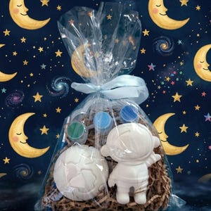 Op de afbeelding: Een cadeauzakje met ruimtethema, met een wit astronautenfiguur en een globe-vormig item. De zak bevat blauwe en groene verfpotjes, alles verpakt in doorzichtig cellofaan en vastgebonden met een blauw lint. De achtergrond heeft een sterrenhemel.