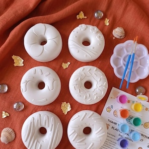 Può includere: Un kit fai-da-te con sei ciambelle di gesso bianco con vari disegni decorativi. Il kit include una tavolozza, pennelli e vasetti di vernice. Le ciambelle sono su un tessuto arancione.