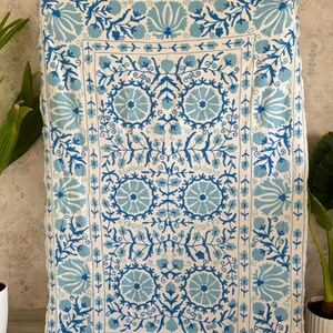 Op de afbeelding: Een rechthoekig textiel wandkleed met een crèmekleurige basis en ingewikkeld blauw bloemenborduurwerk. Het ontwerp bevat een centraal raster van cirkelvormige bloemmotieven omringd door een rand van gestileerde bladeren en ranken in blauw.