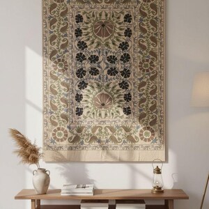 May include: A large, rectangular tapestry with an intricate floral design in beige, brown, green, and blue hues. The tapestry is hanging on a white wall, above a wooden bench with a vase and books.