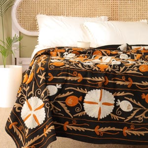 May include: Black embroidered blanket featuring a floral pattern in white, orange, and tan. The blanket is draped over a bed with white pillows and a woven headboard. The detailed design adds a touch of elegance to the bedroom.