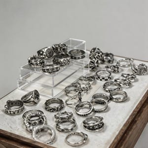 Op de afbeelding: Een verzameling zilveren ringen met diverse ontwerpen, waaronder kruisen, bloempatronen en geometrische vormen. De ringen worden gepresenteerd op een heldere acrylstandaard en een houten oppervlak, waarbij hun gedetailleerde vakmanschap wordt benadrukt.