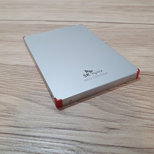 Może przedstawiać: Prostokątny srebrny dysk półprzewodnikowy z czerwonymi akcentami na rogach. Na górnej powierzchni znajduje się czarny nadruk z tekstem SK hynix oraz SOLID STATE DRIVE. Urządzenie posiada gładkie metaliczne wykończenie i smukły profil.