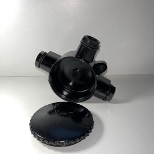 Op de afbeelding: Een zwart fitting met meerdere poorten en een schroefdop. De fitting heeft vier poorten en een centrale opening. De dop is rond met een geribbelde rand. De objecten zijn gemaakt van glanzend zwart materiaal.