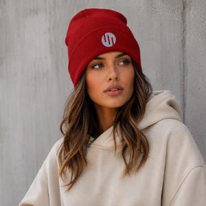May include: A vibrant red beanie featuring a prominent circular logo on the front cuff. The logo consists of a silver-toned graphic design enclosed within a circle. The beanie is constructed with a thick, ribbed knit texture and a wide folded brim.