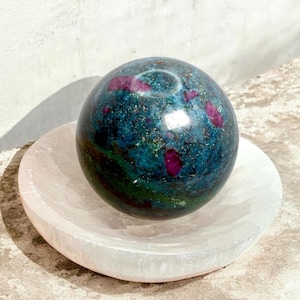 May include: A polished gemstone sphere with a teal and green base color and red inclusions, resting on a white, carved, circular dish. The sphere is approximately 3 inches in diameter. The dish is made of a light pink stone.
