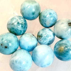 May include: Ten polished, spherical larimar gemstones in varying shades of blue and white marbling. The gemstones are arranged together, showcasing their natural beauty. The blue hues range from light turquoise to deeper shades.
