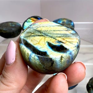 May include: A polished, oval-shaped labradorite stone with iridescent blue, green, and yellow hues. The stone is held in a hand, with other similar stones visible in the background. The labradorite has a smooth, reflective surface.