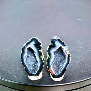 May include: Two matching geode slices with a dark blue and white interior and a black and green outer edge. The geode slices are displayed on a dark grey surface. The text "Set J" is visible at the bottom.