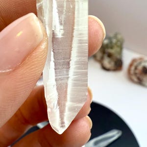 Pode incluir: Um pingente de cristal transparente e alongado com uma argola prateada. O cristal tem uma parte superior áspera e natural e uma parte inferior pontiaguda. O pingente é segurado pelos dedos de uma pessoa. Outros cristais são visíveis no fundo.