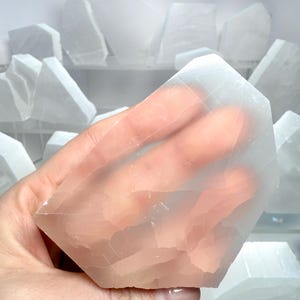 Pu&ograve; includere: Un cristallo di selenite traslucido e sfaccettato &egrave; tenuto in mano, mostrando il suo aspetto naturale e chiaro. Altri pezzi di selenite sono visibili sullo sfondo. La superficie del cristallo presenta striature visibili e una luce morbida e sottile.