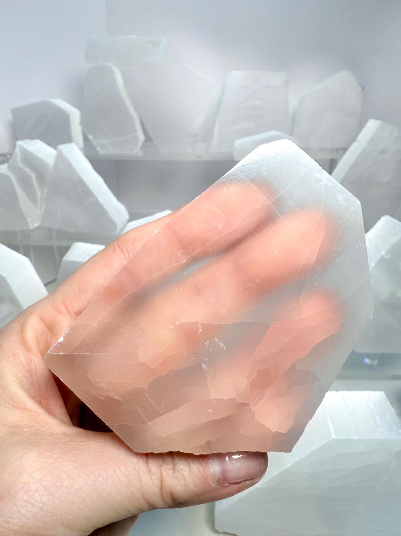 Pu&ograve; includere: Un cristallo di selenite traslucido e sfaccettato &egrave; tenuto in mano, mostrando il suo aspetto naturale e chiaro. Altri pezzi di selenite sono visibili sullo sfondo. La superficie del cristallo presenta striature visibili e una luce morbida e sottile.