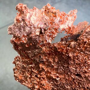 May include: Close-up of a raw copper specimen with a rough, textured surface. The copper has a rich, reddish-brown color with areas of lighter, reflective tones. The irregular shape suggests a natural, unrefined form.