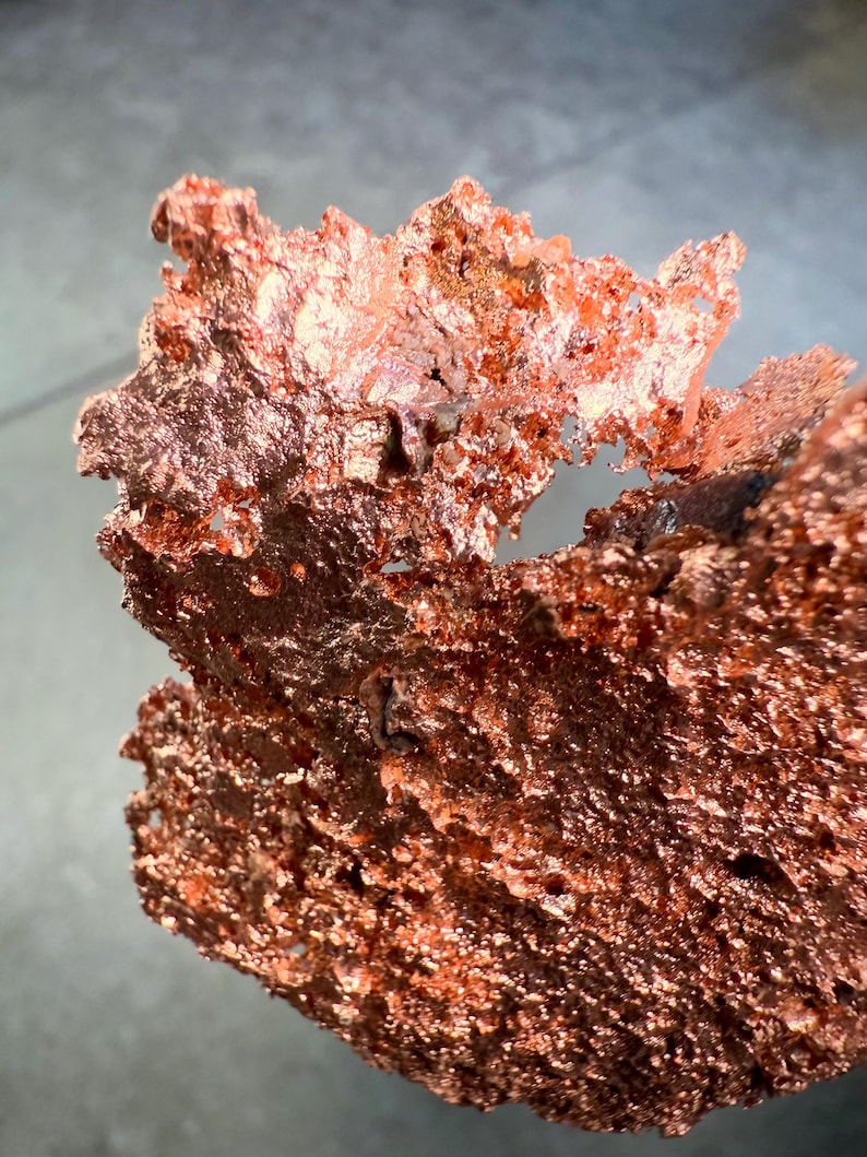 May include: Close-up of a raw copper specimen with a rough, textured surface. The copper has a rich, reddish-brown color with areas of lighter, reflective tones. The irregular shape suggests a natural, unrefined form.