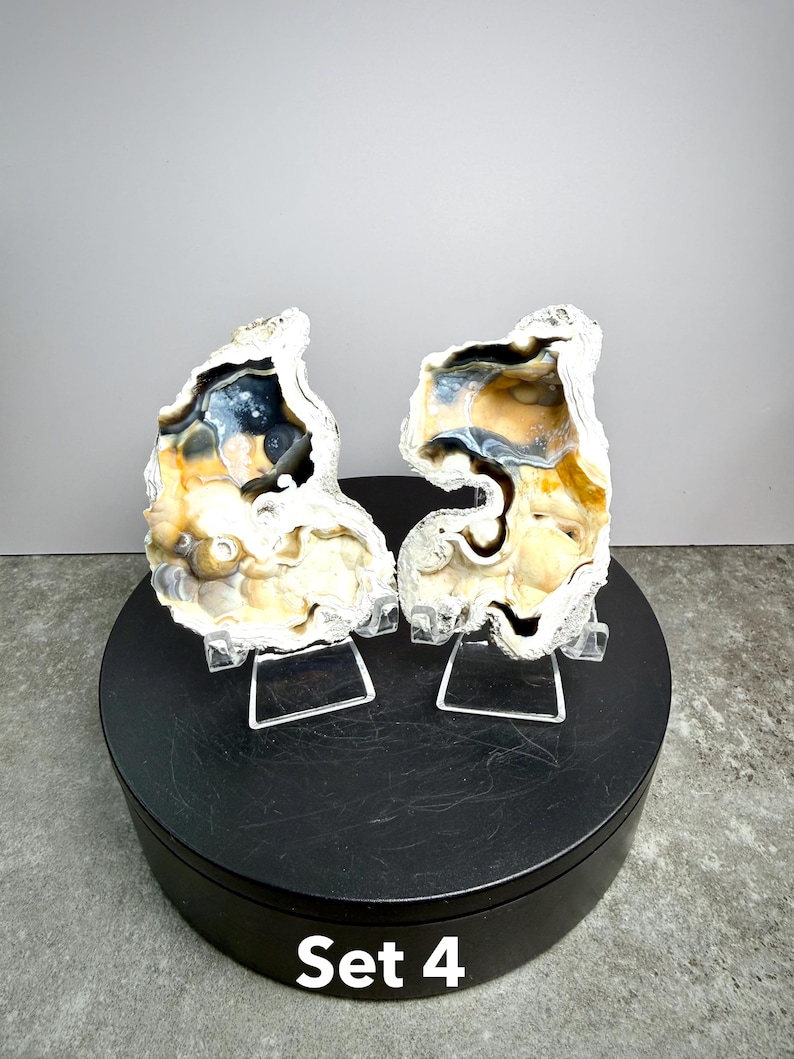 May include: Two geode halves with a mix of colors, including cream, tan, blue, and black, are displayed on clear stands. The geodes are set on a black circular base. The text "Set 4" is visible on the base.
