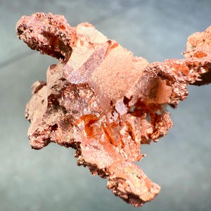 May include: A close-up of a raw copper specimen with a rough, textured surface. The copper has a reddish-brown color with areas of lighter, almost crystalline formations. The specimen is irregularly shaped, with jagged edges and a natural, organic appearance.