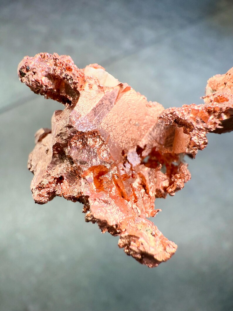 May include: A close-up of a raw copper specimen with a rough, textured surface. The copper has a reddish-brown color with areas of lighter, almost crystalline formations. The specimen is irregularly shaped, with jagged edges and a natural, organic appearance.