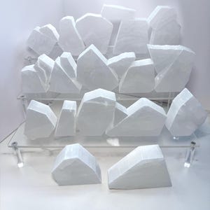 Puede incluir: Una colección de trozos de cristal de selenita blancos, exhibidos en una estantería de acrílico transparente. Los cristales tienen una textura natural rugosa y formas geométricas variadas. La disposición se encuentra sobre un fondo blanco, resaltando la pureza de los cristales.