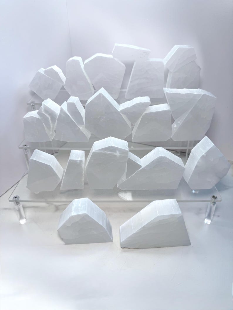 Pu&ograve; includere: Una collezione di frammenti di cristallo di selenite bianchi, esposti su una mensola trasparente in acrilico. I cristalli hanno una consistenza naturale ruvida e varie forme geometriche. La disposizione &egrave; su uno sfondo bianco, evidenziando la purezza dei cristalli.