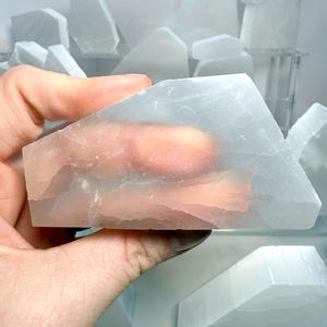 Pu&ograve; includere: Una mano che tiene un cristallo di selenite traslucido, azzurro pallido. Il cristallo &egrave; rettangolare con bordi angolati. Altri cristalli di selenite di varie forme e dimensioni sono sullo sfondo, alcuni su una mensola. I cristalli hanno un aspetto morbido e luminoso.