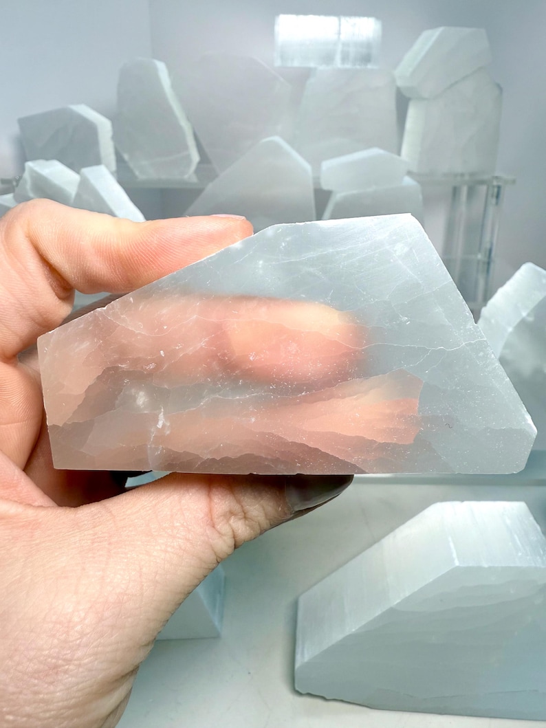 Pu&ograve; includere: Una mano che tiene un cristallo di selenite traslucido, azzurro pallido. Il cristallo &egrave; rettangolare con bordi angolati. Altri cristalli di selenite di varie forme e dimensioni sono sullo sfondo, alcuni su una mensola. I cristalli hanno un aspetto morbido e luminoso.