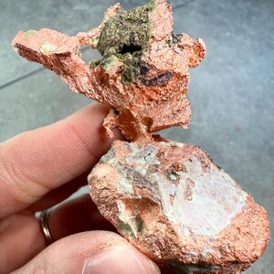 May include: A natural copper specimen with a rough, textured surface. The copper has a reddish-brown hue, with areas of green and dark brown. The specimen is held in a hand, showcasing its irregular shape and mineral composition.