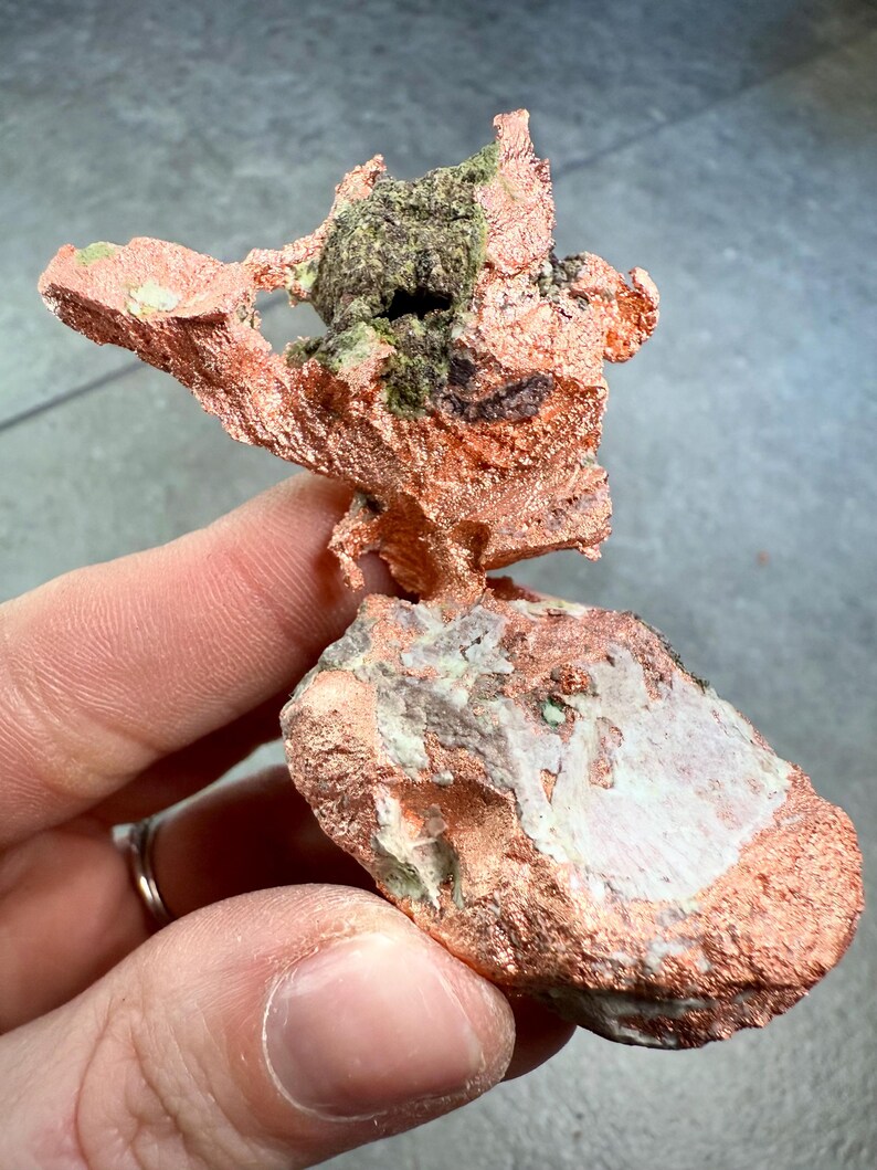 May include: A natural copper specimen with a rough, textured surface. The copper has a reddish-brown hue, with areas of green and dark brown. The specimen is held in a hand, showcasing its irregular shape and mineral composition.