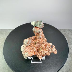 May include: A natural copper specimen with a rough, textured surface. The copper displays shades of reddish-brown and green, and is displayed on a black circular stand. The specimen is supported by a clear acrylic stand.