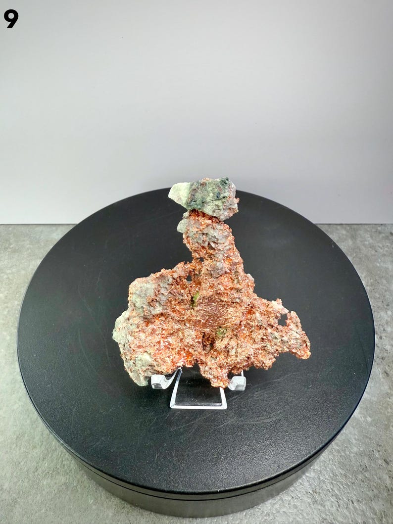 May include: A natural copper specimen with a rough, textured surface. The copper displays shades of reddish-brown and green, and is displayed on a black circular stand. The specimen is supported by a clear acrylic stand.