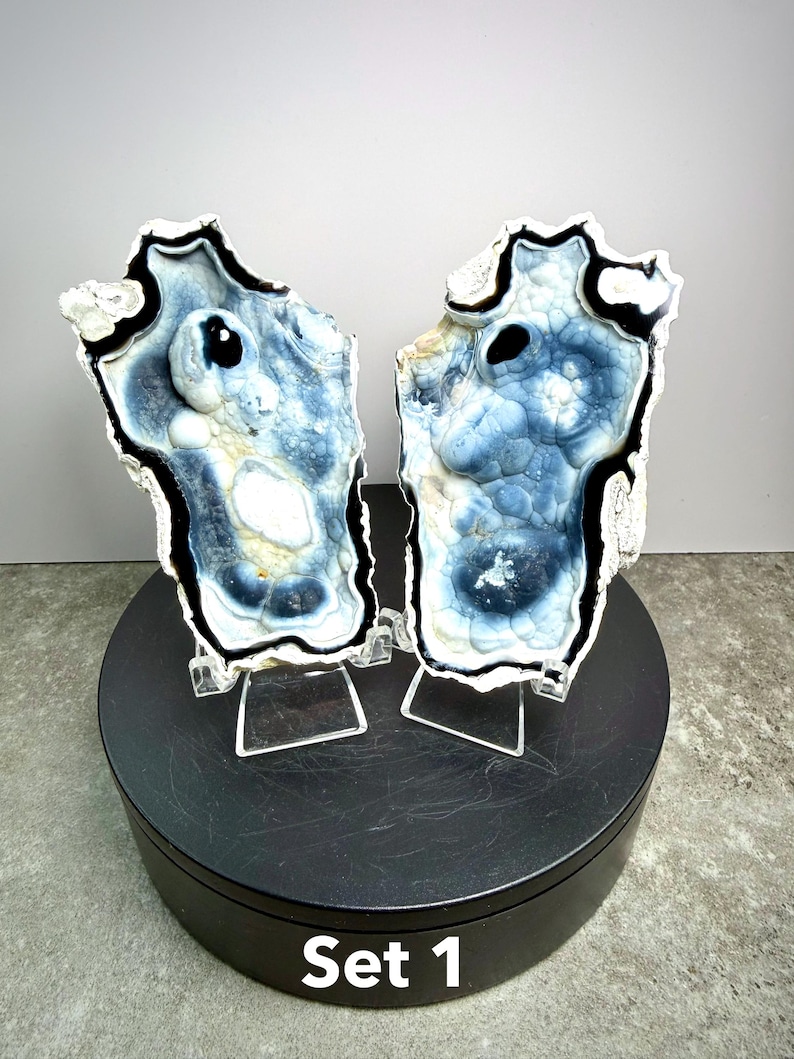 May include: Two matching, irregularly shaped, polished stone slabs with blue, white, and black patterns. The stones are displayed on clear stands atop a black rotating platform. The text "Set 1" is visible on the platform.