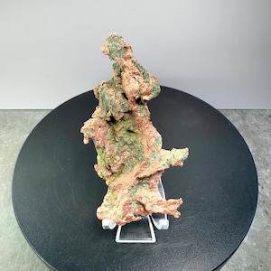 May include: A natural copper ore specimen with a rough, textured surface. The mineral displays a mix of reddish-brown and green hues, mounted on a clear acrylic stand. The specimen is displayed on a black rotating platform.