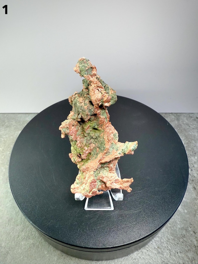 May include: A natural copper ore specimen with a rough, textured surface. The mineral displays a mix of reddish-brown and green hues, mounted on a clear acrylic stand. The specimen is displayed on a black rotating platform.
