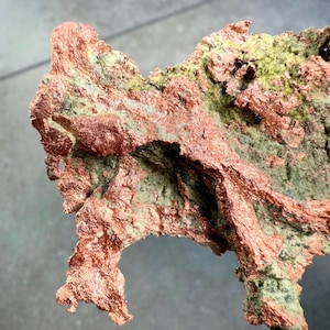 May include: A close-up of a copper mineral specimen with a rough, textured surface. The copper has a reddish-brown color with patches of green and gray hues. The mineral's shape is irregular and jagged, with a natural, organic appearance.