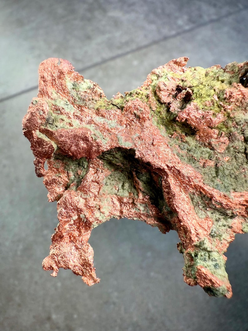 May include: A close-up of a copper mineral specimen with a rough, textured surface. The copper has a reddish-brown color with patches of green and gray hues. The mineral's shape is irregular and jagged, with a natural, organic appearance.