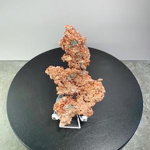 May include: A raw copper specimen with a rough, textured surface. The copper has a reddish-brown color with hints of green and is displayed on a clear stand. The specimen is set against a black circular base and a neutral background.