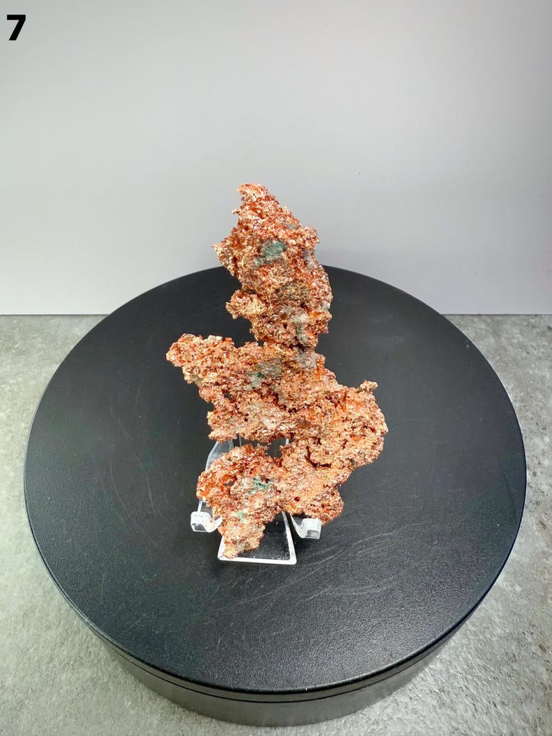 May include: A raw copper specimen with a rough, textured surface. The copper has a reddish-brown color with hints of green and is displayed on a clear stand. The specimen is set against a black circular base and a neutral background.