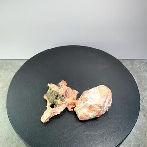 May include: Two raw mineral specimens. One is a reddish-brown, irregularly shaped piece with a green patch. The other is a light orange, rounded rock with white patches. Both are displayed on a black circular surface.
