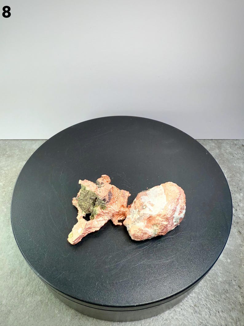 May include: Two raw mineral specimens. One is a reddish-brown, irregularly shaped piece with a green patch. The other is a light orange, rounded rock with white patches. Both are displayed on a black circular surface.