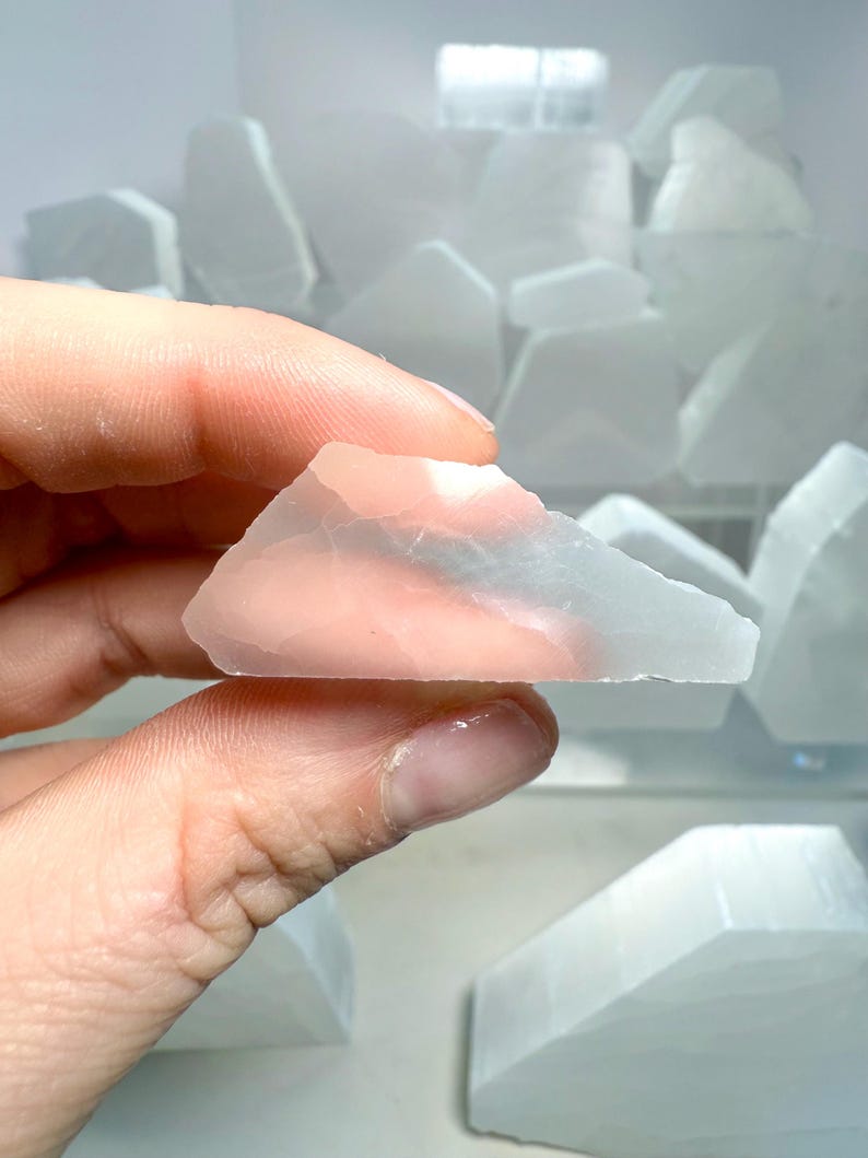 Pu&ograve; includere: Un cristallo di selenite traslucido rosa chiaro tenuto in mano, con una superficie ruvida e irregolare. Altri cristalli di selenite sono visibili sullo sfondo, di varie dimensioni e forme.