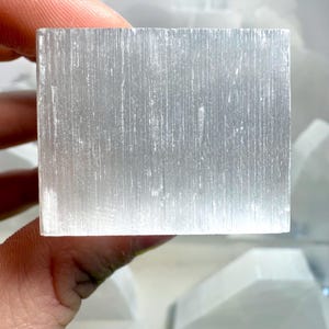 Pu&ograve; includere: Un cristallo di selenite rettangolare traslucido. Il cristallo &egrave; di un colore bianco-argento pallido con striature verticali visibili. Altri pezzi di selenite sono visibili sullo sfondo. Il cristallo &egrave; tenuto in una mano.