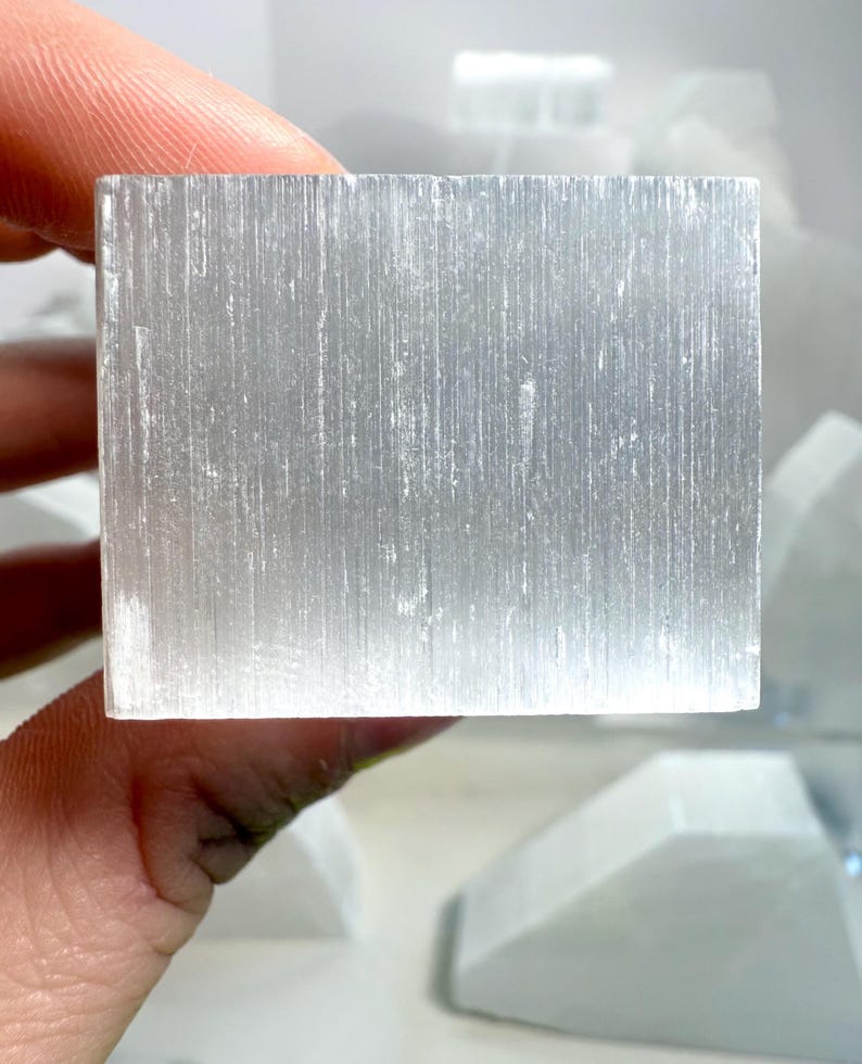 Pu&ograve; includere: Un cristallo di selenite rettangolare traslucido. Il cristallo &egrave; di un colore bianco-argento pallido con striature verticali visibili. Altri pezzi di selenite sono visibili sullo sfondo. Il cristallo &egrave; tenuto in una mano.