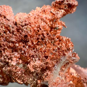 May include: Close-up of a raw copper specimen. The copper has a rough, textured surface with a reddish-brown color. The specimen has a crystalline structure with a porous appearance. The background is a soft gray.